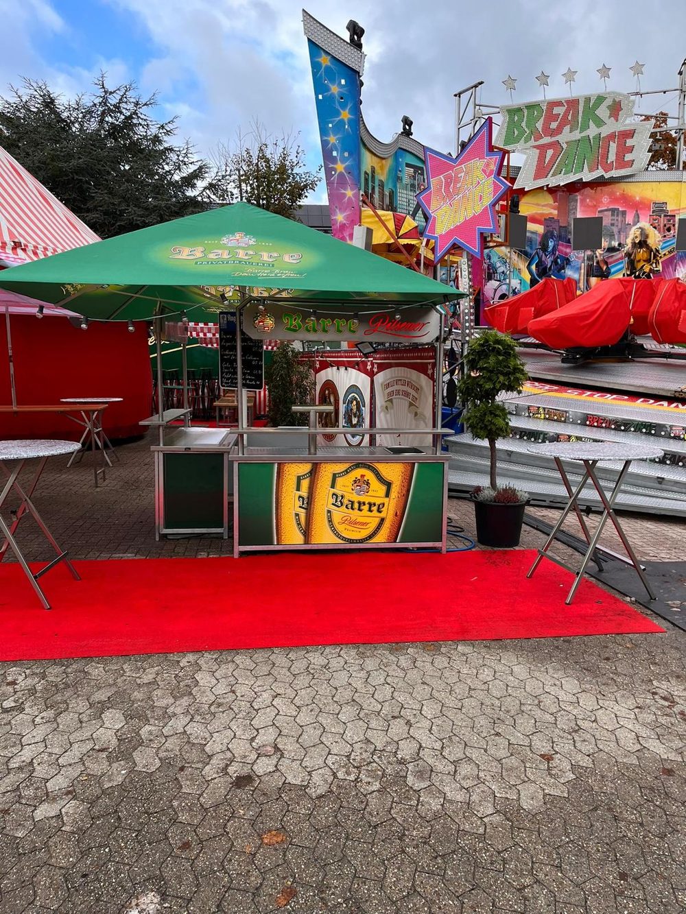 Ausschank mit grünem Barre-Schirm auf der Kirmes, im Hintergrund das Fahrgeschäft Break Dance