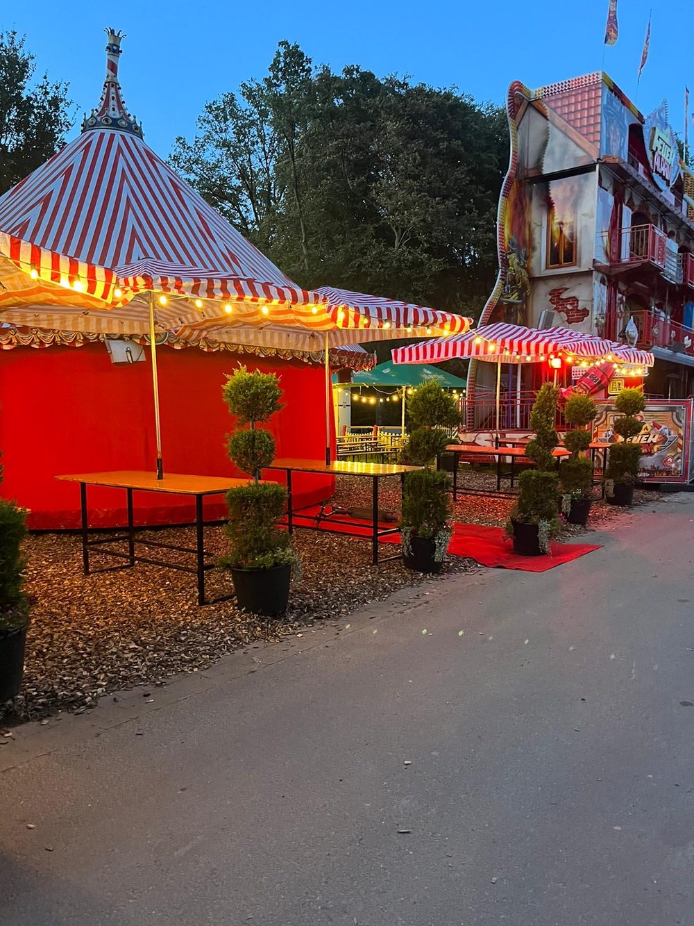 Rot-weiß gestreiftes Zelt und Tische mit Lichterkette in der Dämmerung, daneben ein Fahrgeschäft